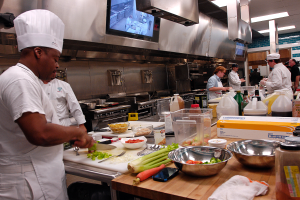 Eine Gruppe von Köchen in einer Küche, die auf einem Schneidbrett Essen zubereiten, mit einem Koch im Vordergrund in einer weißen Schürze und Mütze, der ein Messer hält und von Utensilien, Schüsseln, Gemüse, einem Handy, Flaschen und anderen Gegenständen umgeben ist, mit einem Fernseher und Deckenleuchten im Hintergrund.