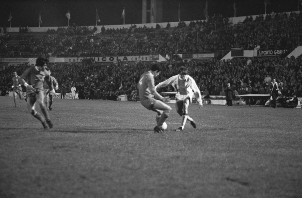 Schwarzes und weißes Foto von Männern, die Fußball auf einem Feld in einem Stadion spielen, mit Zuschauern auf den Tribünen und Fahnen im Hintergrund.