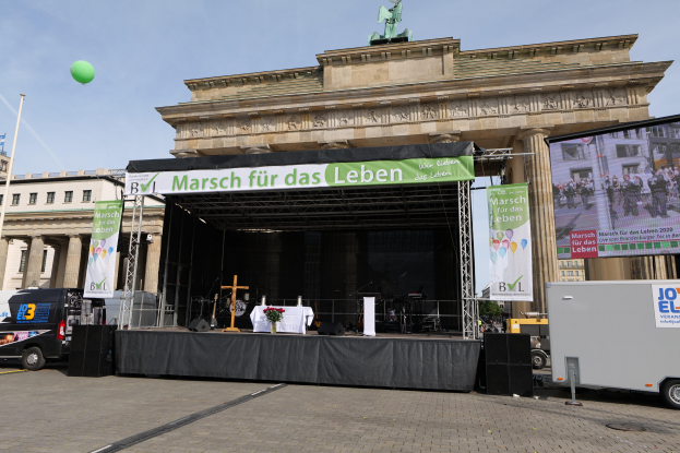 Eine Bühne vor dem Brandenburger Tor in Berlin, Deutschland, mit einem Tisch mit einer Blumenvase, einem Kreuzsymbol, Transparenten, Lautsprechern und anderen Gegenständen, sowie Fahrzeugen auf der Straße und Gebäuden im Hintergrund.