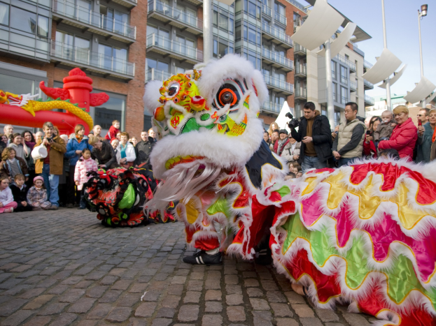 Ein lebendiges chinesisches Neujahrsfest in Amsterdam mit einer Löwen-Tanz-Show vor einer Menschenmenge, einige halten Kameras, vor Gebäuden und Laternen unter einem klaren blauen Himmel.