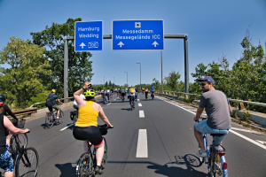 Gruppe von Radfahrern mit Helmen, die eine Straße mit einem Geländer und Bäumen auf beiden Seiten entlangfahren, Lichtmasten im Hintergrund, unter einem klaren blauen Himmel, mit einem Schild, das eine Radtour in Hamburg anzeigt.