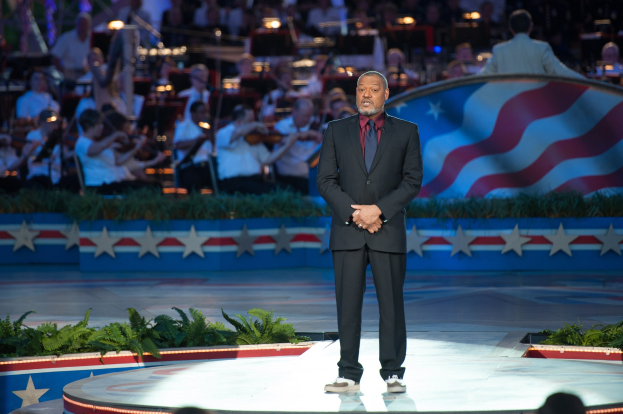 Will Smith in Anzug und Krawatte auf einer Bühne vor einem Publikum mit Musikinstrumenten haltenden Menschen, mit Pflanzen und einer Flagge im Hintergrund während einer Debatte.