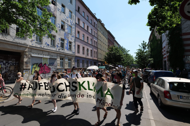 Eine Gruppe von Menschen marschiert auf einer Straße und hält ein Banner hoch, mit parkenden Autos auf der rechten Seite, einem Fahrradfahrer auf der linken Seite und Gebäuden, Bäumen und einem Schild im Hintergrund unter einem sichtbaren Himmel.