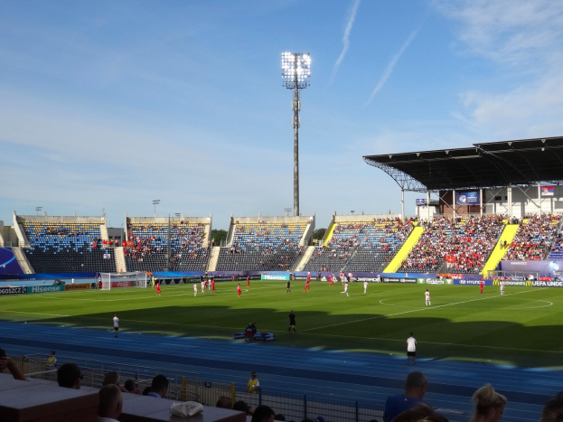 Ein Fußballspiel in einem großen Stadion mit Spielern auf dem Feld und Zuschauern in den Rängen unter Flutlicht, mit sichtbarem Himmel im Hintergrund.