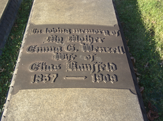 Grabstein mit der Inschrift "Die liebevolle Erinnerung an meine Mutter" in einem grasbewachsenen Friedhof mit welken Blättern.