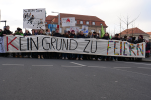 Demonstranten mit einem Banner mit der Aufschrift "Kein Grund zu Feiern" gegen deutsche Sparmaßnahmen, mit Straßeninfrastruktur und Gebäuden im Hintergrund.