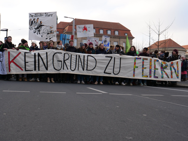 Demonstranten mit einem Banner mit der Aufschrift "Kein Grund zu Feiern" gegen deutsche Sparmaßnahmen, mit Straßeninfrastruktur und Gebäuden im Hintergrund.