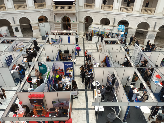Große Besuchermenge auf einer internationalen Fachmesse mit Ständen, Bannern, Tischen, Stühlen und Luftballons in einem Raum mit Bogengängen, Säulen und einem Bildschirm im Hintergrund.