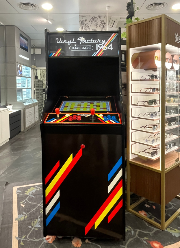 Ein Geschäftsinnenraum mit einem zentralen Retro-Arcade-Automaten, einem Glasvitrinenschrank auf der rechten Seite und einer retro-stilvollen Beleuchtung mit Schränken und einem Fernseher im Hintergrund.