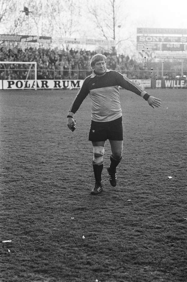 Ein professioneller Fußballspieler in Fußballuniform steht auf einem Feld umgeben von Zuschauern, einem Torpfosten und Bäumen unter einem klaren Himmel.