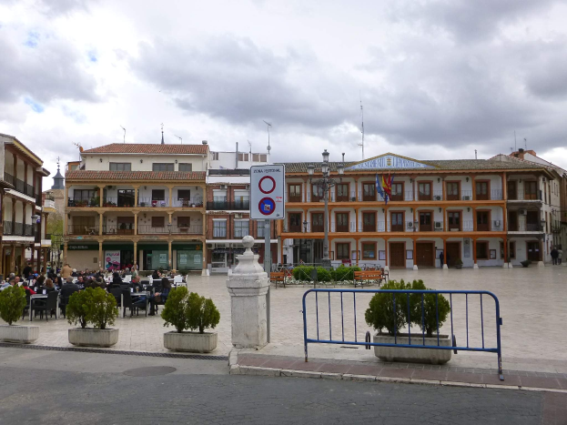 Ein belebter Stadtplatz mit sitzenden und stehenden Menschen, Topfpflanzen, Metallabsperrungen, einem Schild an einem Pfahl, Straßenlaternen mit Fahnen, umliegenden Gebäuden mit Fenstern und einem bewölkten Himmel.