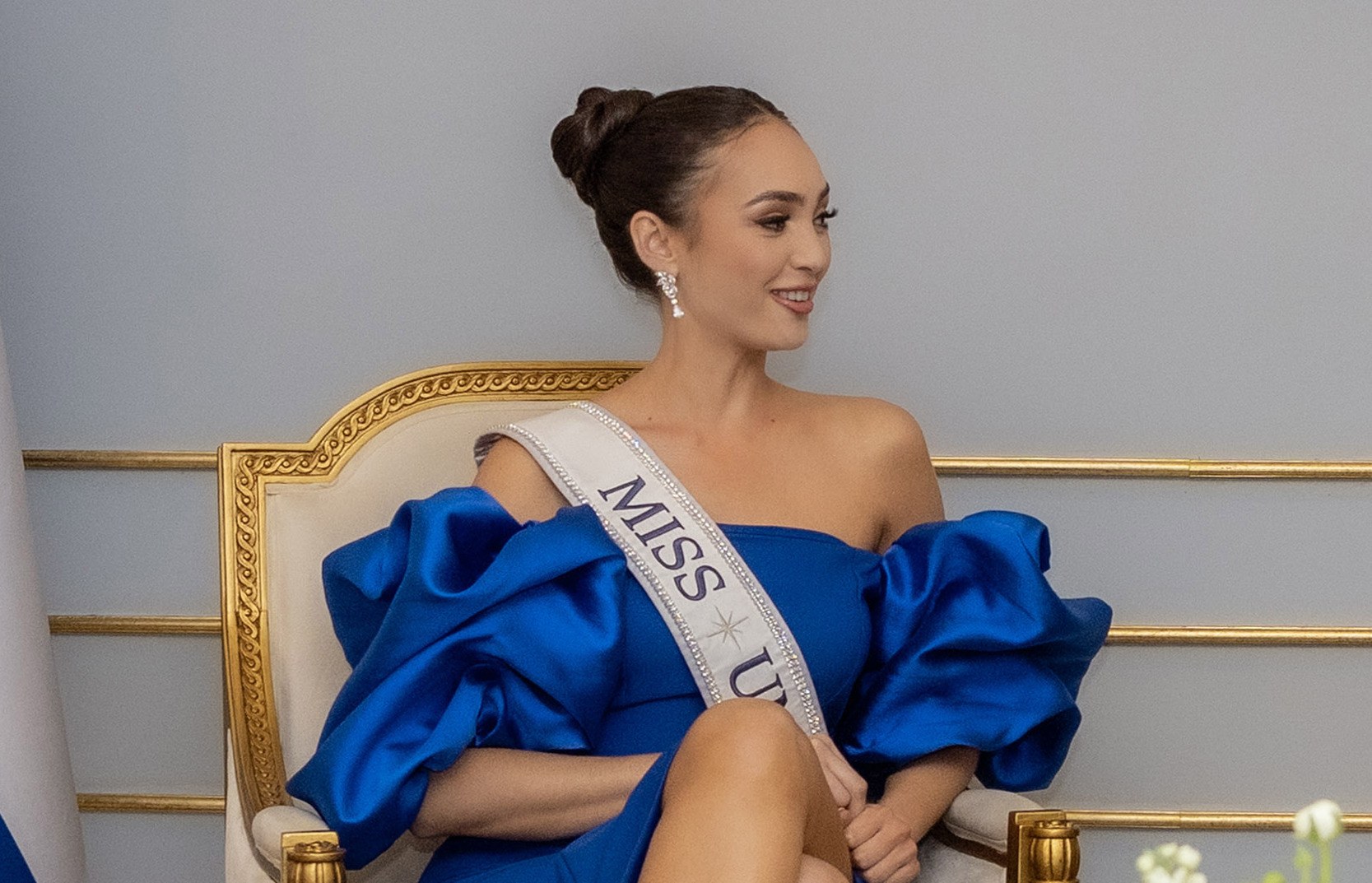 Frau in einem blauen Kleid mit einem "Miss Universe 2019" Band, sitzend auf einem Stuhl umgeben von Blumen mit einem Lächeln.