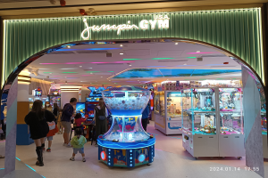 Eine Gruppe von Menschen steht vor dem Eingang einer Sporthalle mit einem Bogen, einem Anzeigebildschirm und Deckenleuchten, einige halten Taschen, mit einem Wasserzeichen unten.