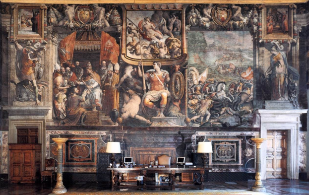 Großer Raum mit einem großen Gemälde an der Wand, ein Tisch mit Gegenständen, zwei Lampen, zwei Türen und ein Teppich, befindet sich im Palazzo della Signoria in Florenz, Italien.