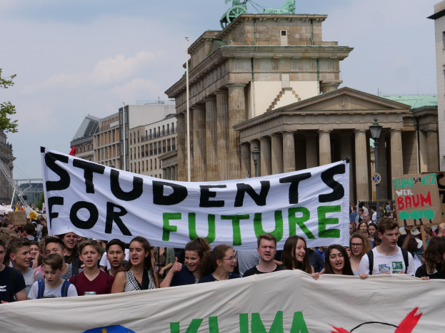 Gruppe von Schülern marschiert in Berlin mit einem buntfarbenen "Students for Future"-Schild gegen eine Kulisse aus Gebäuden, Bäumen und Himmel.