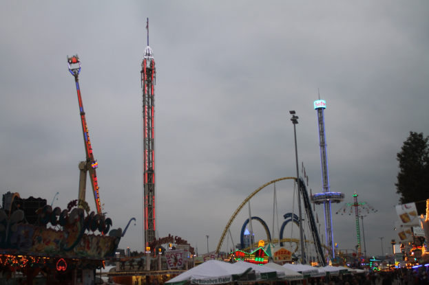 Eine Menschenmenge bei einer nächtlichen Karnevalsveranstaltung, beleuchtet von bunten Fahrgeschäften, Pfählen und Bäumen unter einem bewölkten Himmel.