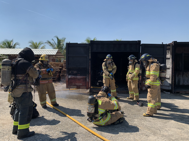 Feuerwehrleute in Helmen und Ausrüstung vor einem Gebäude mit Rohren stehend, mit Bäumen, Schuppen und hölzernen Gegenständen im Hintergrund während einer scheinbar Feuerlöschtrainingssitzung.