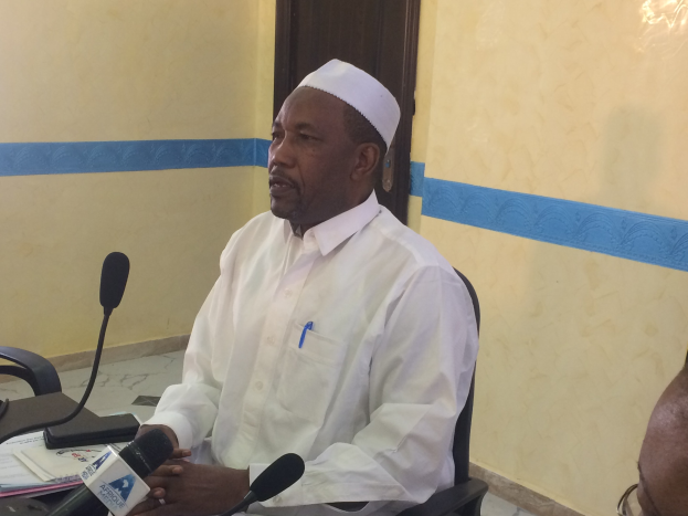 Ein Mann in einem weißen Kleid und Hut sitzt an einem Tisch mit Mikrofonen, spricht, während Papiere und Gegenstände auf dem Tisch sind, mit einer anderen Person mit Brille rechts von ihm, in einem Raum mit einer Wand und einer Tür im Hintergrund.